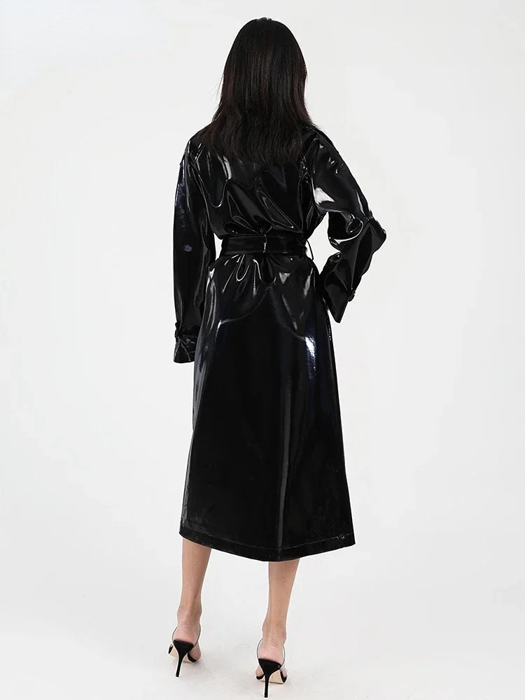 A woman viewed from behind wearing a glossy black long coat or wrap with a tie waist, paired with black high heels, against a plain light background.