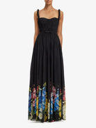 A sleeveless black maxi dress with a pleated bodice, thin shoulder straps, a matching waist tie, and a vibrant multicolor floral print along the lower hem, shown on a model against a plain white background.
