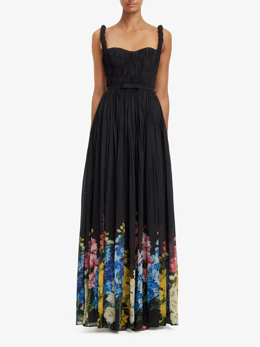 A sleeveless black maxi dress with a pleated bodice, thin shoulder straps, a matching waist tie, and a vibrant multicolor floral print along the lower hem, shown on a model against a plain white background.