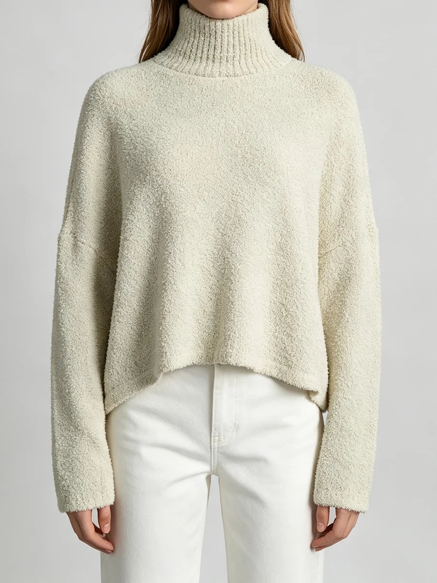 A woman wearing a cream-colored, textured bouclé turtleneck sweater and white pants, shown from the neck down against a light gray studio background.